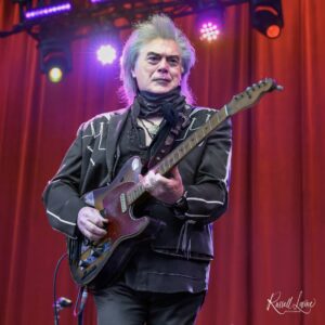 Musician playing an electric guitar on stage, wearing a black outfit and scarf, with red curtains and colorful stage lights in the background.