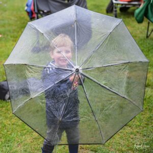 Child standing in a grassy outdoor area holding a large transparent umbrella in the rain.