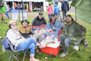 Four friends sit in folding chairs under a canopy at an outdoor festival, sharing snacks from a red cooler and a patchwork blanket in front of them.