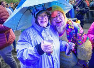 Two people in colorful costumes share a moment under a blue umbrella at a crowded outdoor event, smiling at the camera.