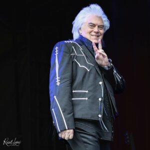 Older man with white hair on stage, wearing a black studded jacket, giving a peace sign and smiling at the camera (signature watermark bottom left).