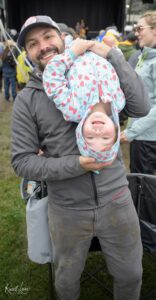 Man in a gray jacket holding a smiling child upside down in a light blue strawberry-patterned outfit at an outdoor event.