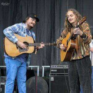 Two musicians playing acoustic guitars on a stage, one in a blue plaid shirt and cap, the other in a striped brown sweater with long hair.