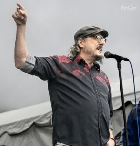 Male performer on stage, wearing a dark red and black patterned shirt, speaking into a microphone with one arm raised in a fist.