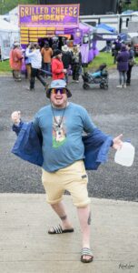 Cheerful man in a blue shirt and bucket hat poses with a jug at an outdoor festival, with a purple grilled cheese booth in the background.