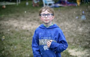 Boy wearing a blue hoodie and glasses stands in a grassy outdoor area with bubbles floating around him.