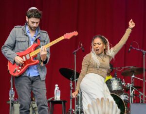 Male guitarist in a gray jacket playing a red electric guitar on stage beside a vocalist with her arm raised against a red curtain backdrop.