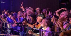 Crowd at a concert raising hands, illuminated by purple and blue stage lights behind a barrier.
