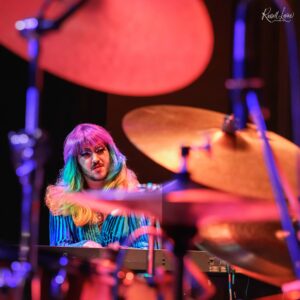 Musician with rainbow hair playing a keyboard on a dimly lit stage bathed in red and blue lights.