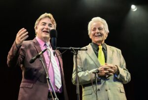 Two male performers in suits on stage, speaking into microphones and smiling at the audience.