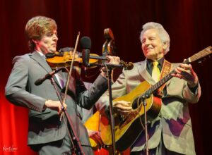 Two musicians in gray suits perform on stage: one plays the violin, the other the guitar, with a red curtain backdrop.