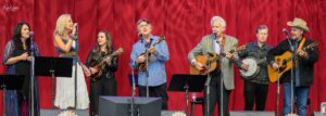 Seven musicians perform on a stage in front of a red curtain, singing and playing guitars and a banjo.
