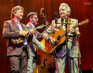 Three male musicians in suits performing on stage with guitars and a microphone.