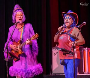Two drag performers on stage: one with purple outfit and purple wig singing into a microphone while playing a guitar; the other in a blue-pink costume with bold makeup sings into a mic.