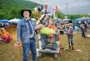 Smiling man in a denim jacket, hat, and striped tie stands beside a cluttered, decorated cart at an outdoor festival.