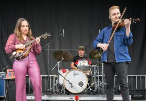 Female mandolin player in a pink outfit performs on stage beside a male violinist in a blue jacket; drummer in back.