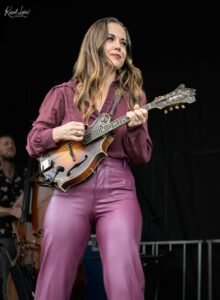 Female musician in a maroon blouse playing a sunburst mandolin on stage with a dark background.