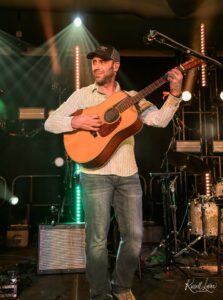 Male guitarist performing on stage with an acoustic guitar under colorful stage lights