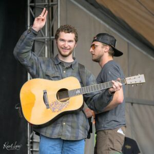 Two male musicians on stage; the left guitarist waves a peace sign while holding an acoustic guitar in front of a dark backdrop.