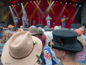 Close-up of two concertgoers wearing hats in the crowd—a beige woven hat with pins on the left and a black top hat with a feather on the right, with a blurred stage in the background.
