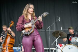Female musician in a burgundy outfit playing a mandolin on stage, with other band members and drums visible in the background.