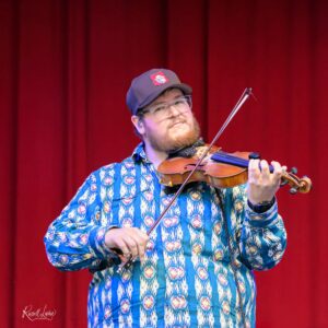 Violinist performing on stage with a red curtain backdrop; wearing a blue geometric shirt and cap.