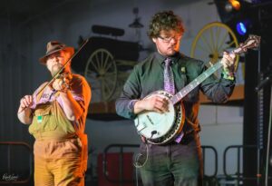 Two male musicians perform on a stage: a banjo player in a dark shirt and a fiddler in overalls beside him, with wagon-wheel decor in the background.