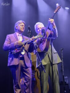 Two male musicians perform on stage with guitars and microphones under purple stage lighting.