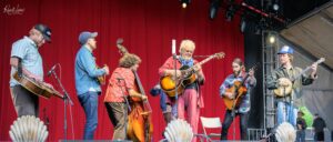 Seven musicians on a stage with a red curtain, playing guitars, a double bass, and a banjo.