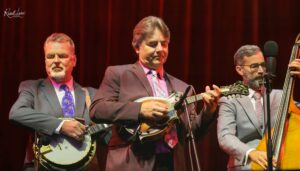 Three male musicians in suits performing on a red-curtained stage, each with a string instrument (banjo, guitar/mandolin, and upright bass).