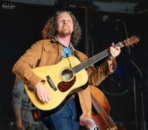Male guitarist performing on stage with an acoustic guitar, wearing a tan jacket.