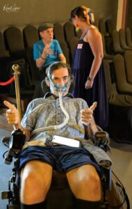 Older man in a hospital gown in a wheelchair wearing an oxygen mask, giving a thumbs‑up while a woman in a navy dress with a rainbow bow stands nearby.