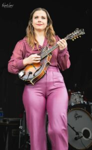 Female musician on stage, playing a sunburst mandolin in a burgundy outfit, with a drum kit visible in the background.