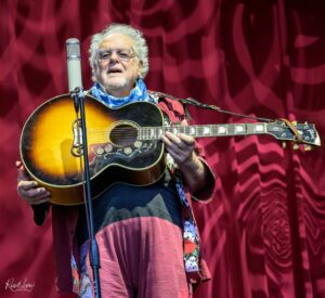 Older man with gray hair and glasses playing an acoustic guitar on stage against a red curtain backdrop.