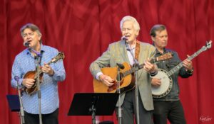 Three male musicians perform on a stage with red curtains: two guitarists flank a lead singer in a gray suit, while a banjo player stands to the right.