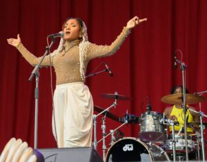 Female singer on stage, arms raised, singing into a microphone with a red curtain backdrop.