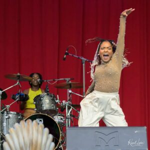 Energetic singer on stage with raised arm, wearing a beige sparkly outfit, performing beside a drummer against red curtains.