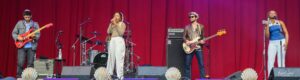 Live band performing on a red-curtain stage: guitarist left, center female lead singer, drummer behind, bassist to the right, and a second female vocalist at far right on mic.