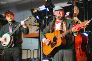 Two male musicians performing on stage: one sings into a microphone while playing an acoustic guitar, wearing a striped cap and red bow tie; the other plays a banjo beside him.