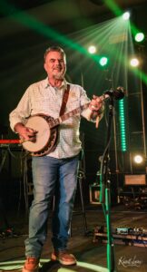 Male musician on stage playing a banjo under green stage lights during a live performance.