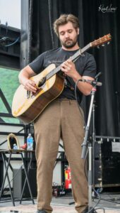 Male guitarist with a beard playing an acoustic guitar on an outdoor stage at a performance, focused on fretting notes.