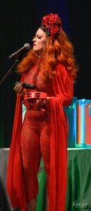 Drag performer in a red rhinestone bodysuit with a large red rose headdress, singing into a microphone and holding a red tambourine on stage.