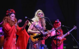 Female singers on stage performing live; two women in red outfits with floral headpieces sing into microphones while playing guitars.