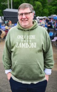 Smiling man wearing a green hoodie that reads 'ONLY HERE FOR THE ZAMBONI' at an outdoor event.