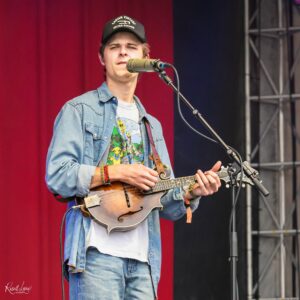 Male musician performing on stage with a mandolin, wearing a denim shirt and cap, singing into a microphone against a red backdrop.