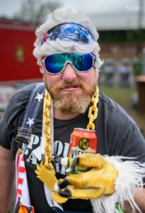 Bearded man with white hair wearing a patriotic bandana and blue mirrored goggles, with a chunky yellow chain around his neck and a red can visible at chest level.