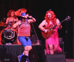 Three performers on a dim stage in colorful outfits: a drummer left, a central dancer in a star-spangled hat and blue mesh outfit, and a pink-dressed guitarist on the right.