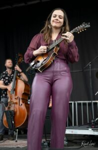 Female musician on stage playing a mandolin, wearing a maroon blouse and pants, with a bassist in the background on a dark stage.