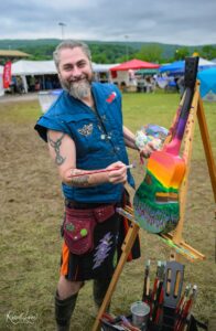 Bearded man in a blue vest paints a rainbow guitar sculpture on an easel at an outdoor art market.