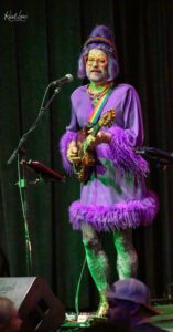 Performer in a bright purple outfit with feather trim playing an electric guitar on stage, wearing a purple wig, red glasses, and colorful necklace at a microphone stand.